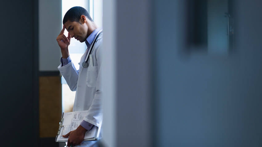 Doctor leaning against desk with clipboard in dark room with anxiety