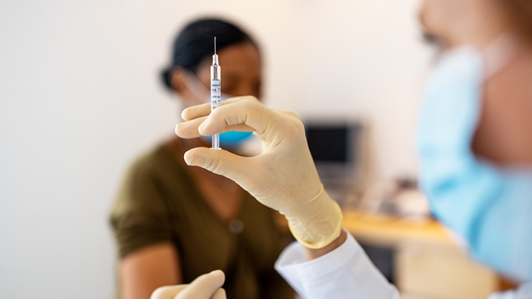 health worker holding shot in focus with mask on with patient with mask sitting in background with mask on