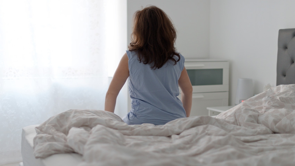 woman sitting on bed blue shirt white sheets wall