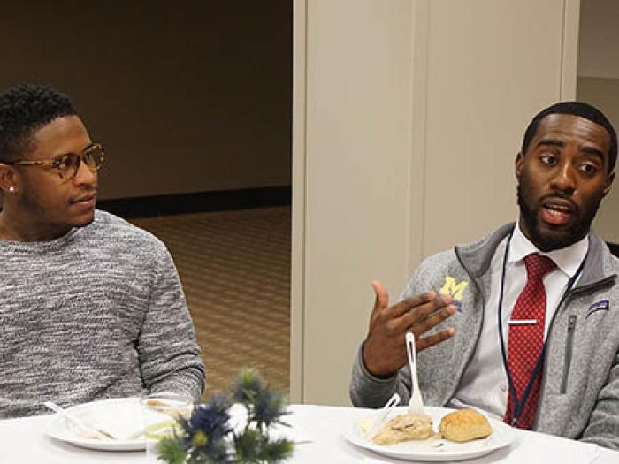 Two black men seated at a table with a white tablecloth and plates of food with one speaking