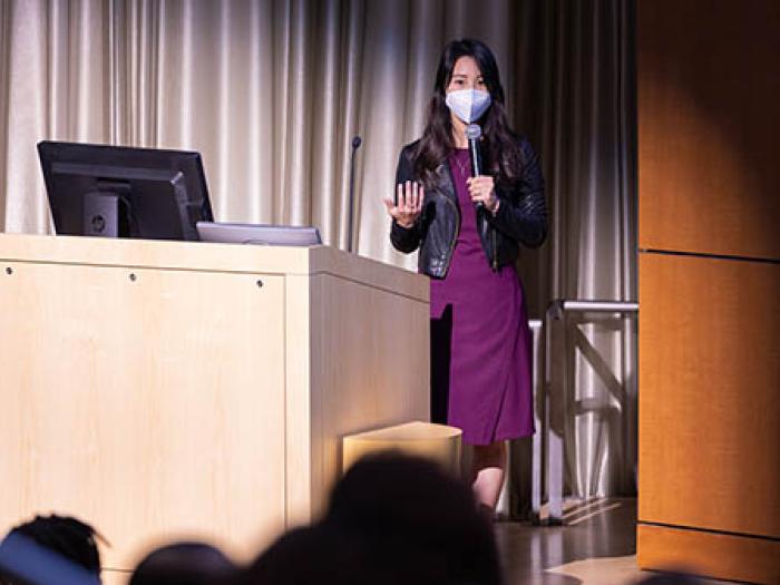 Asian woman wearing mask, purple dress and black leather jacket standing next to a podium on a stage