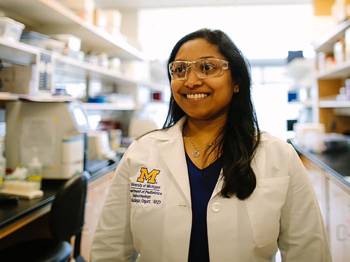 Female doctor with brown skin and dark hair wearing white coat with block M and surgical goggles in lab