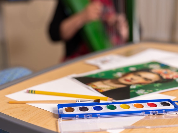 A tray of watercolors sits on a table next to papers and a portrait