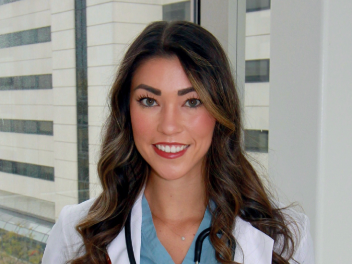 A young Asian American woman with long curled brown hair, wearing blue scrubs, a white doctor's coat, and a stethoscope