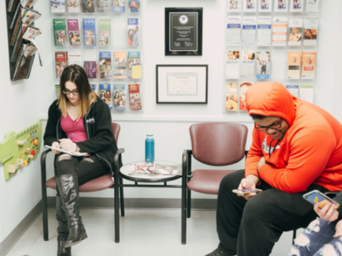A group of young people sitting in the waiting room of a clinic. Some are on their phones, some are filling out paperwork on a clipboard.