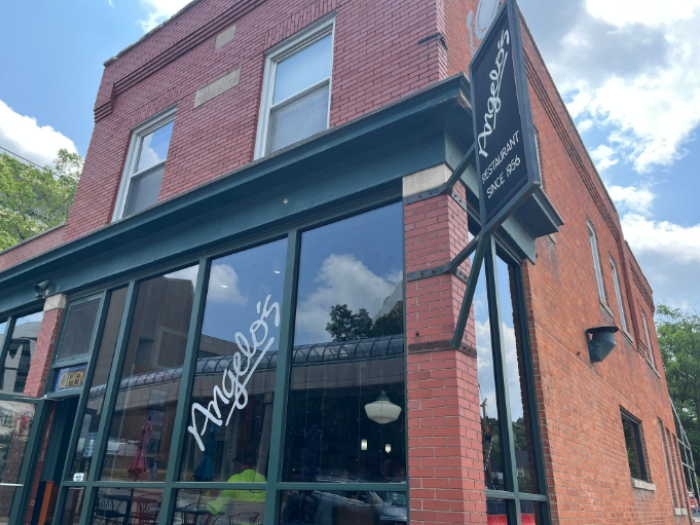 A brick building with "Angelo's" written on the window. It's a summer day, with blue sky reflected in the windows.