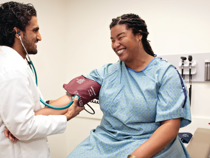 A medical student in a white coat takes the blood pressure of a standardized patient (medical actor) who is wearing a medical gown and sitting on an exam table. They are both smiling.