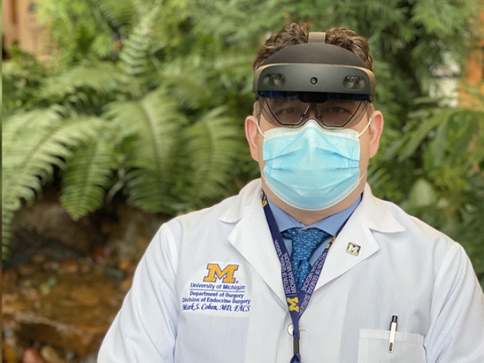 doctor outside in white coat with headset on and blue mask on in front of greenery