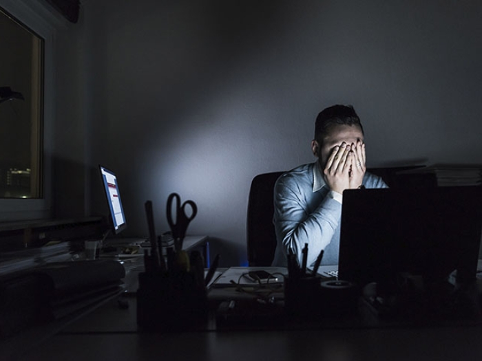 Man sitting behind a computer in a darkened room with his hands over his face