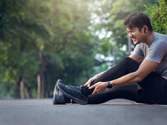 Man sitting on sidewalk among green trees holding his hurt ankle