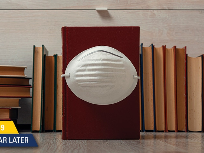 mask on brown books with covid anniversary stamp on bottom left in yellow and blue