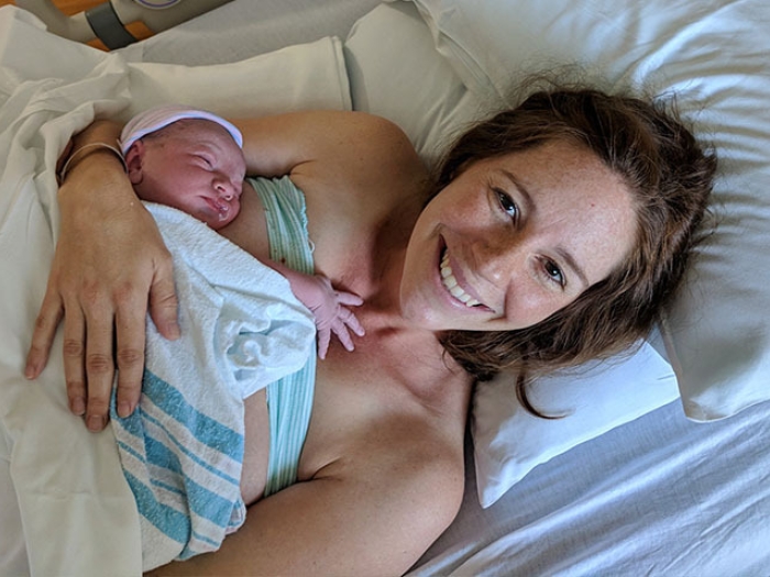 Kate laying in white hospital bed with white sheets holding son with blue and pink hat on after delivery