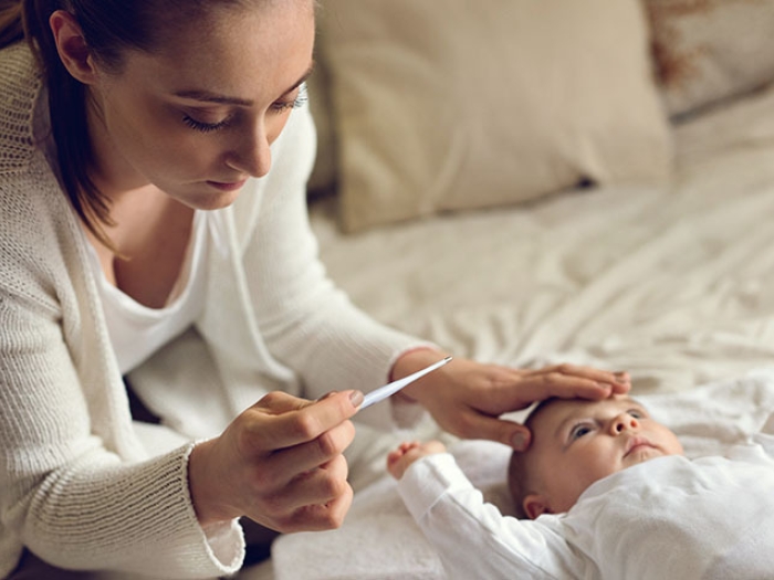 Mom on bed with white sheets wearing white with baby feeling head and looking at thermometer in hand.