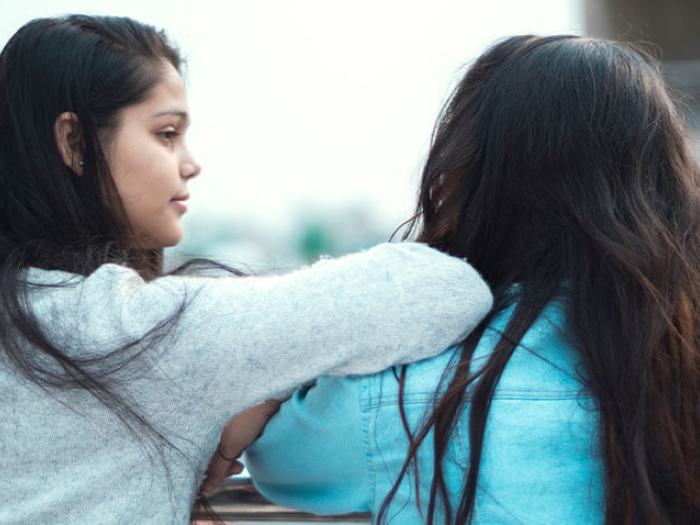 girl with long black hair looking and leaning on friend's shoulder with long brown hair and blue shirt