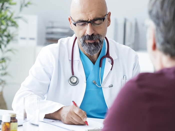 doctor with white coat sitting with stethoscope around his neck and blue shirt talking with patient with short grey hair and maroon shirt