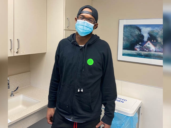 man standing in doctors office with mask on and black coat and glasses