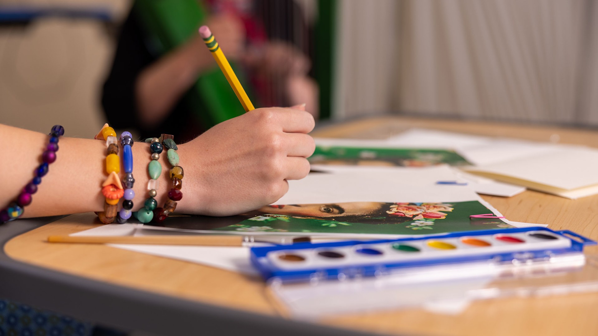 Woman's hand holding pencial with colorful bracelets on wrist and art supplies on table