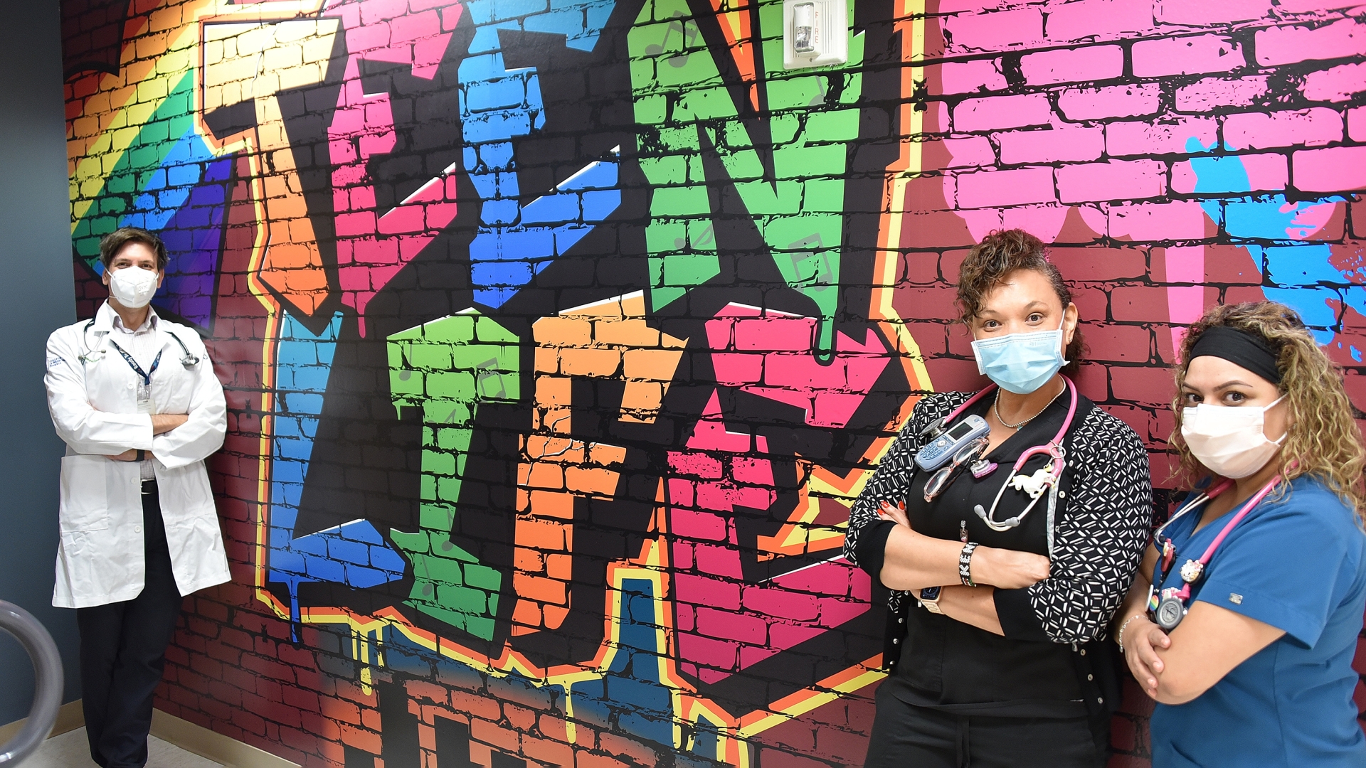Multi-colored words "Teen Life" painted on brick wall with healthcare professional and teens standing with arms folded in front
