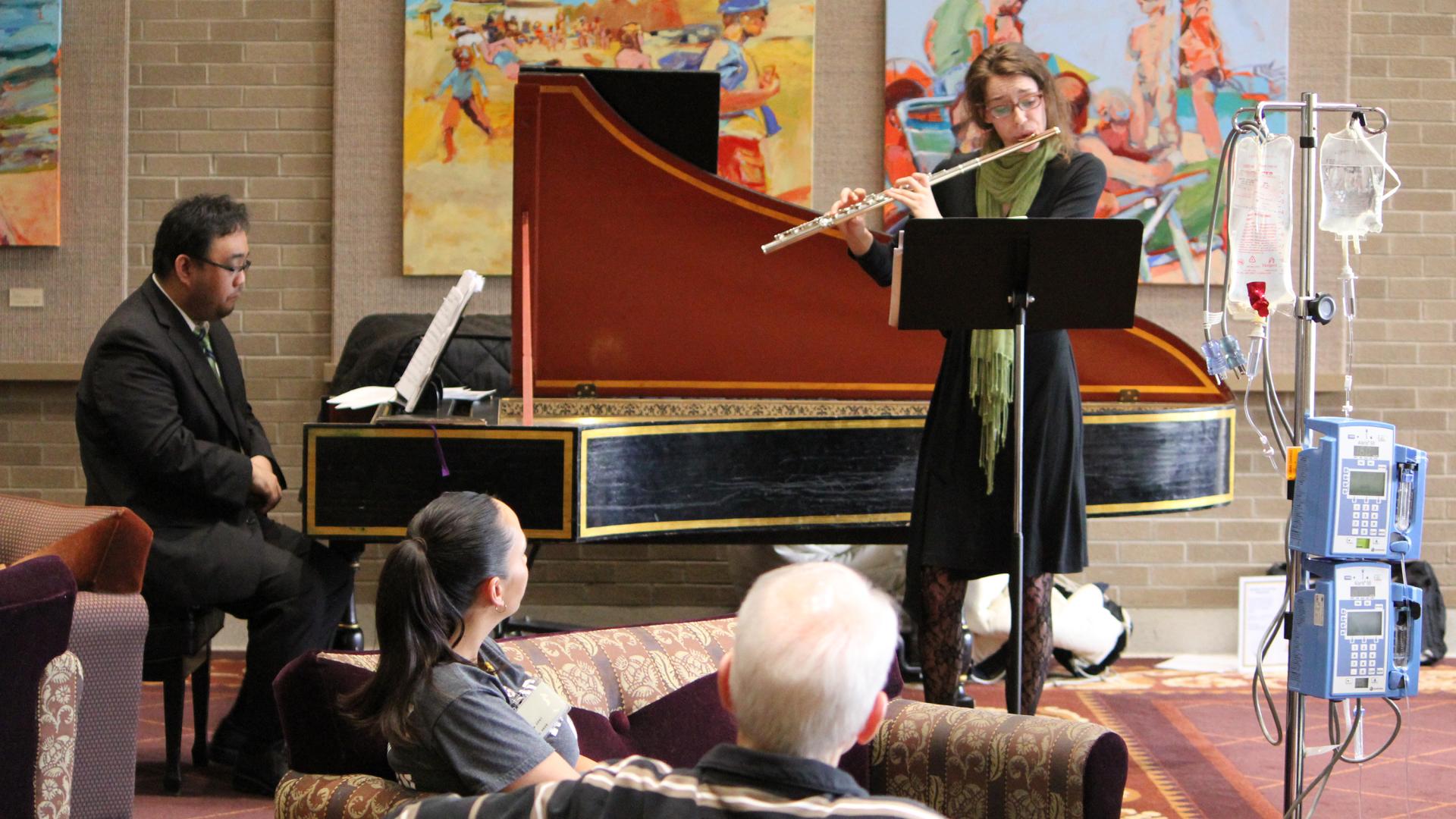 Woman playing flute for patients with artwork hanging on brick wall behind and IV pole with medical equipment in foreground
