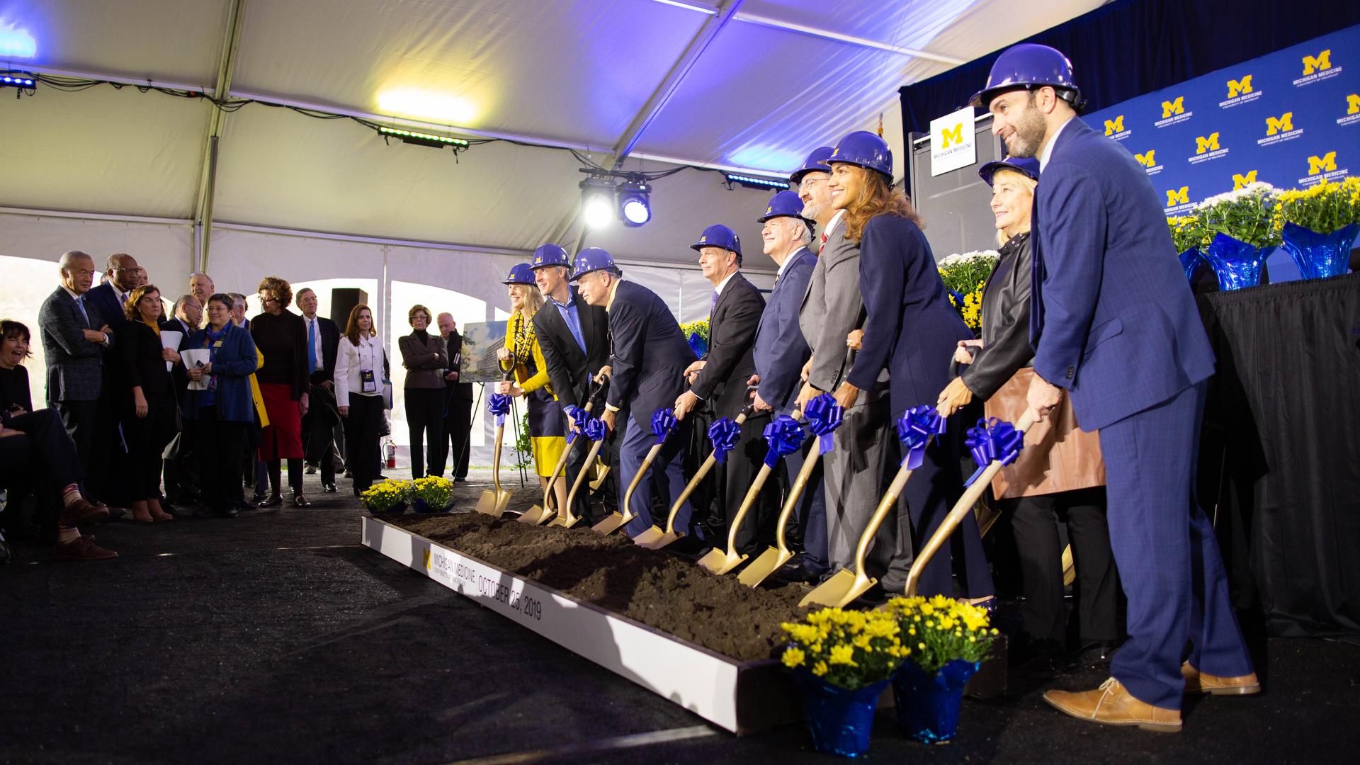 Row of leaders wearing construction helmets with shovels in dirt and Michigan block M background