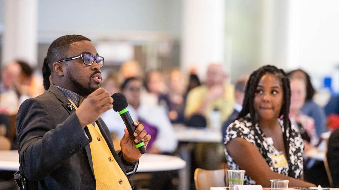 Black man wearing glasses, yellow vest and suit coat, speaking with microphone while seated at table