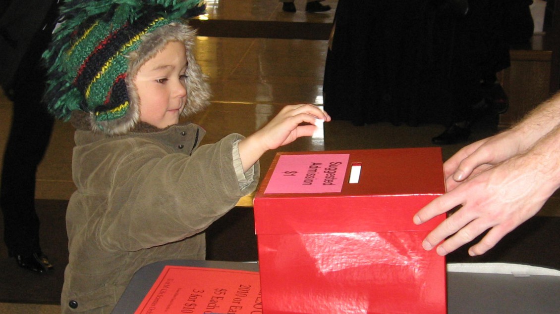 Little boy with striped green knit hat with earflaps putting something in a red donation box