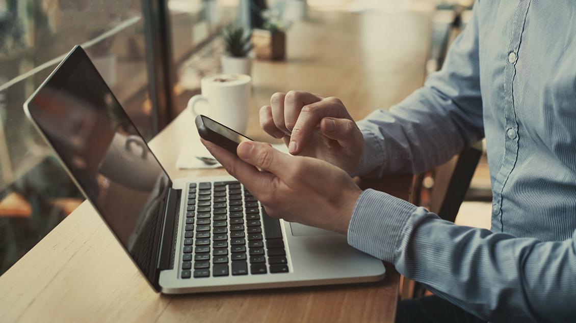 man in blue button-down shirt holding a mobile phone in front of a laptop