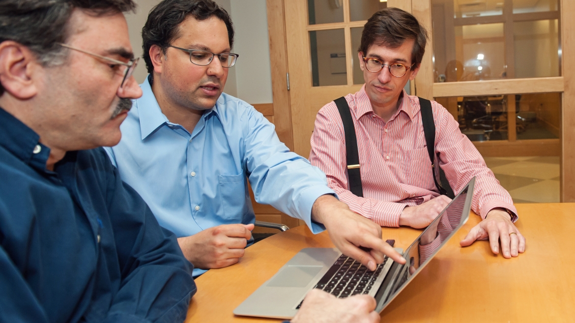 Three men looking at a laptop; one person pointing at the screen.