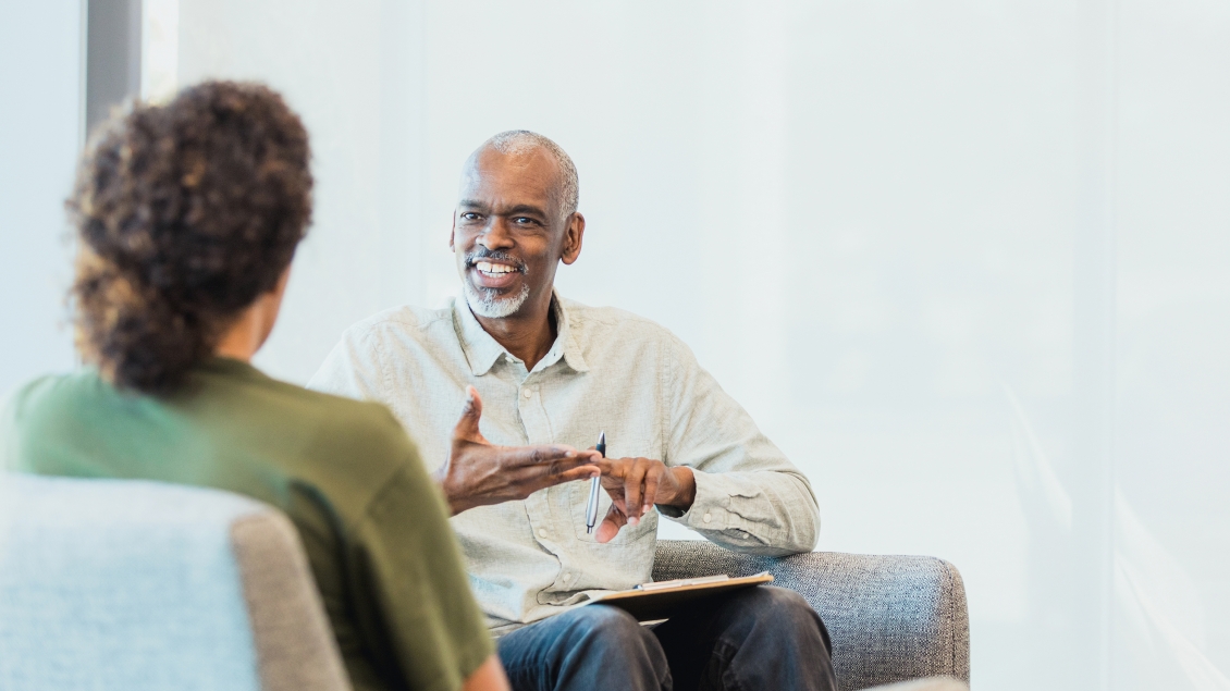 An unrecognizable mid adult woman listens as the cheerful mature adult male counselor gestures and speaks.