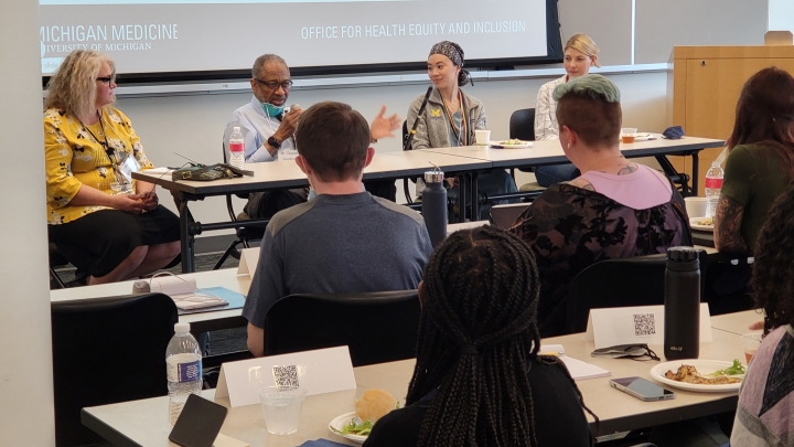 Black man gesturing and seated with group at table talking to students with back to camera
