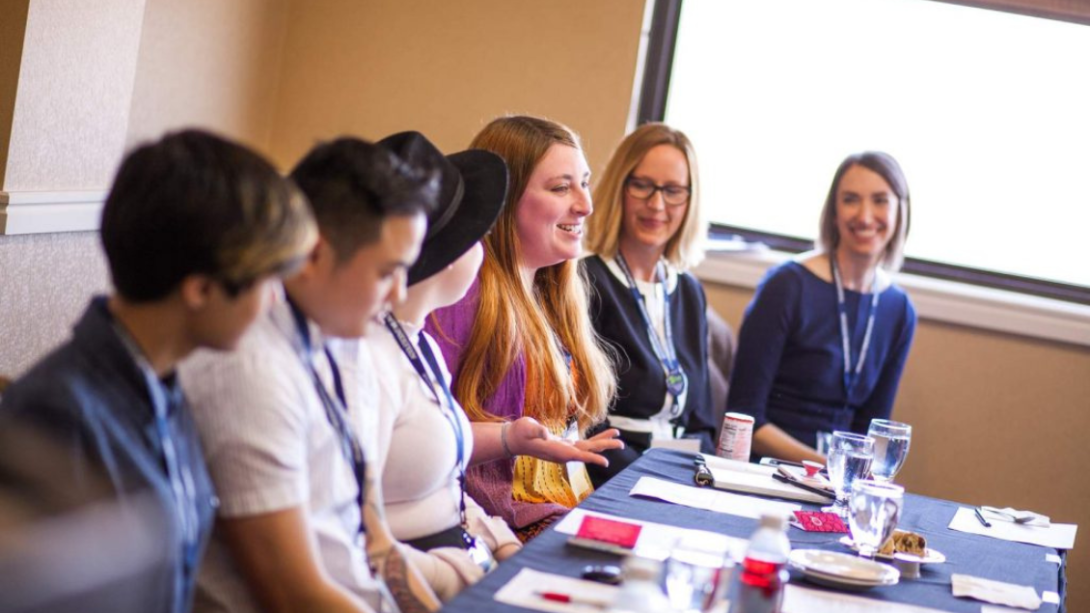 Group of teens sitting on one side of a table wearing name badges and smiling while one person talks