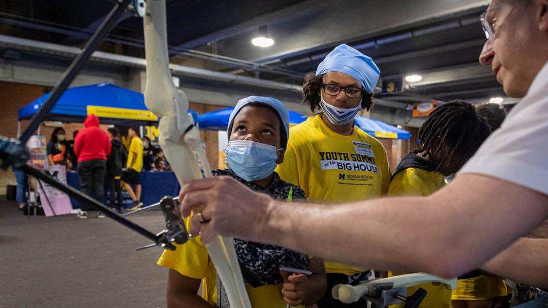 Man with glasses showing leg-bone function to group of Black boys and young men wearing masks and maize t-shirts with text: Youth Summit at the Big House