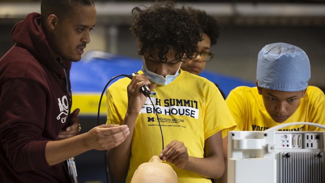 Young black males with maize Youth Summit at the Big House t-shirts watching as one works with medical equipment