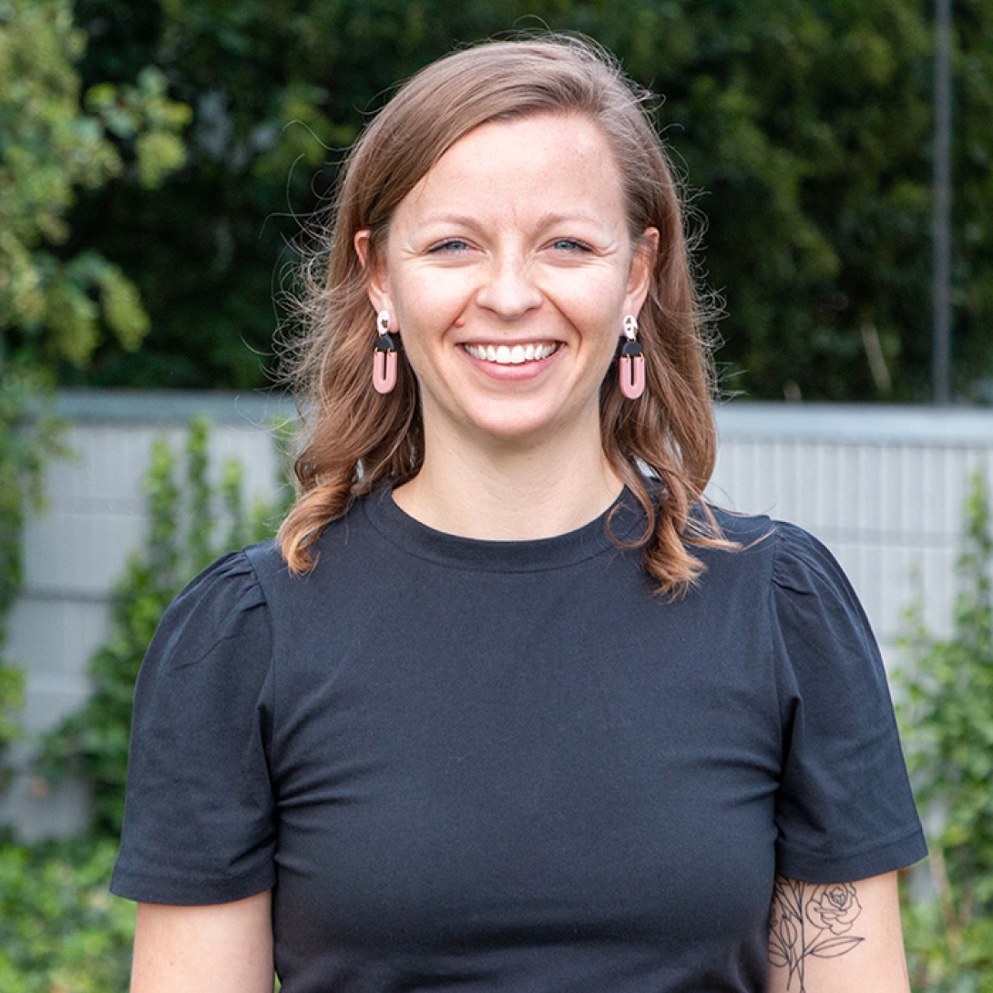 Smiling white woman standing outside with light brown hair wearing black dress and pink and black hoop earrings